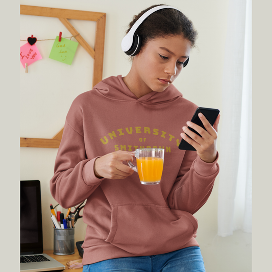 Young woman wearing a Heritage brown “University of Smithdown” hoodie, holding a glass of orange juice and looking at her phone while sitting by a desk.