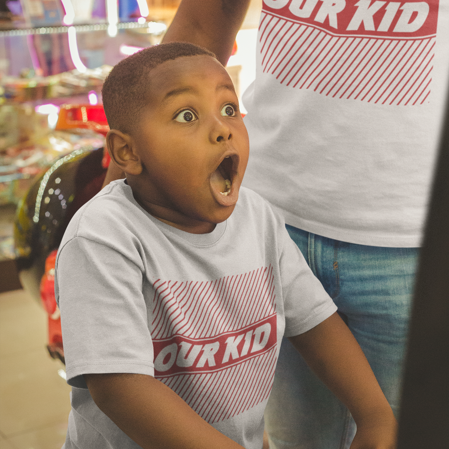 Young boy with a shocked expression wearing a natural raw coloured “Our Kid” t-shirt with red graphic stripes, standing next to an adult wearing an adult version of the t-shirt, both indoors near arcade lights.
