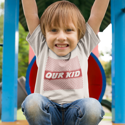 Happy child wearing a  “Our Kid” t-shirt with red graphic stripes, playing on a playground and smiling brightly.