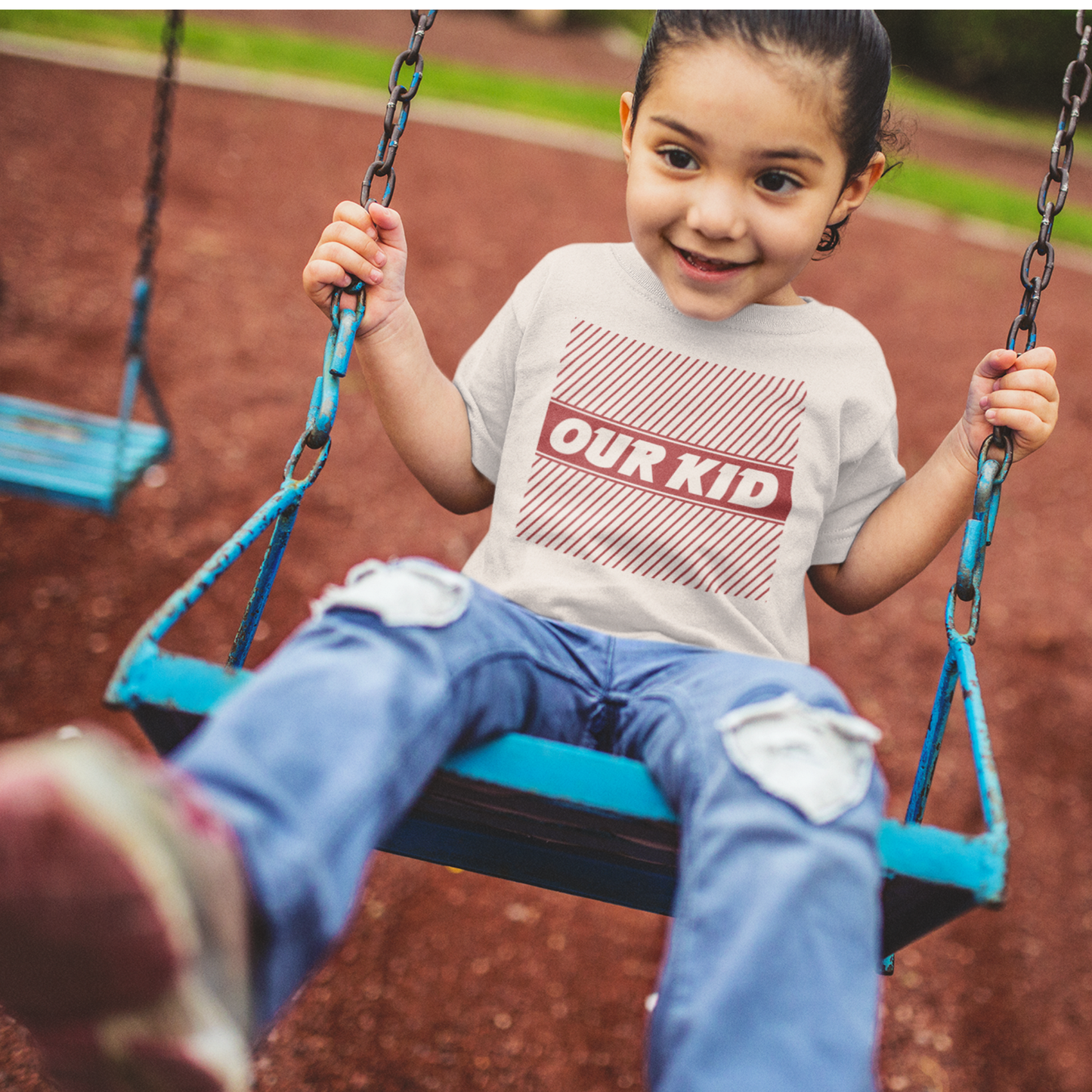 Child on a swing wearing a shirt with 'OUR KID' text  with red graphic stripes in a playground setting