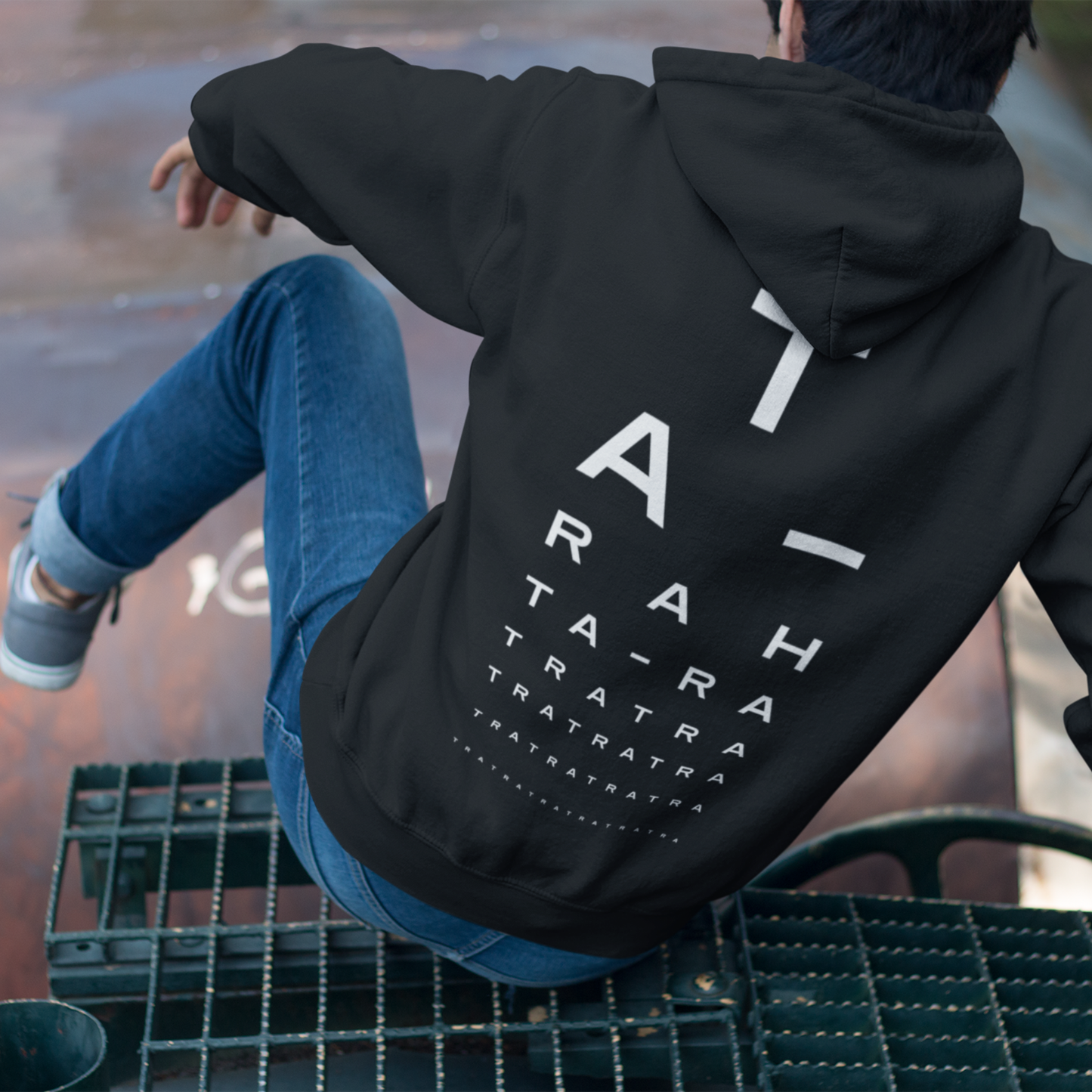 Person sitting on a metal structure wearing a black hoodie with bold white “TA-RA” graphic text on the back, styled like an eye chart.