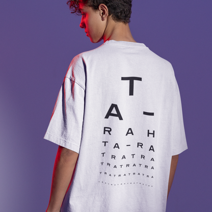 Person wearing a white oversized t-shirt with black “TA-RA” typographic design on the back, styled like an eye test, against a purple-lit background.
