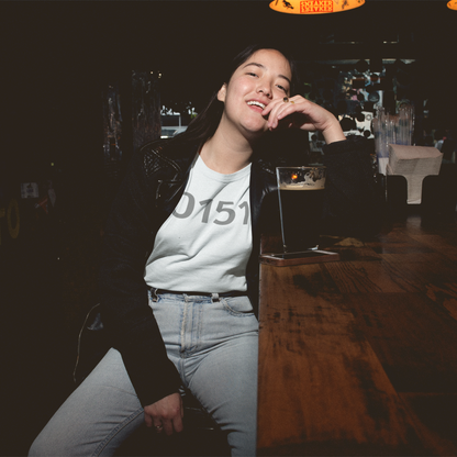 Woman wearing a grey “0151” t-shirt, sitting at a bar table with a pint of stout, smiling casually.
