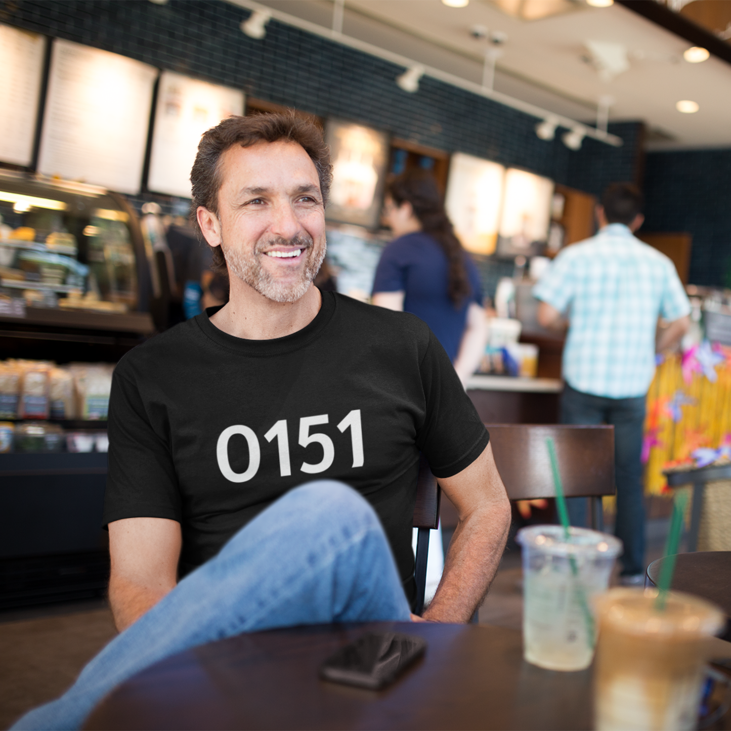 Man wearing a black “0151” t-shirt, sitting in a café with a coffee, smiling and relaxed.