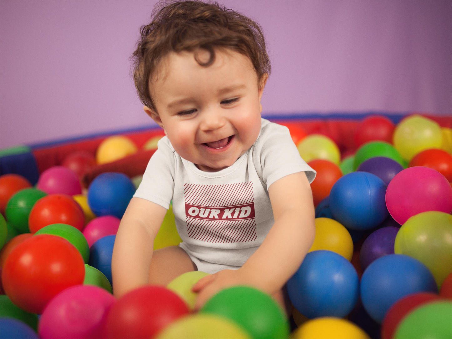 Child in a ball pit wearing a 'Our Kid' onesie with a purple background